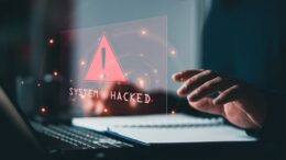 Cyber security warning displayed on a laptop screen with a red alert icon and the message 'System Hacked', highlighting the cost of a cyber attack or security breach.
